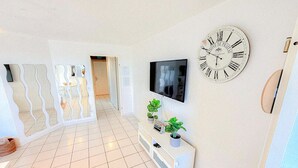 Un salon moderne avec une grande horloge murale, un téléviseur à écran plat et une console blanche agrémentée de plantes.