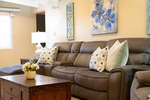 A living room with a brown sofa, patterned cushions, a wooden coffee table with a vase of flowers, and a wall with floral artwork.