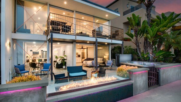 Una moderna casa a due piani con un balcone, un'area salotto esterna e un elemento decorativo a fuoco.