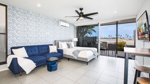 Una moderna camera d'albergo con un divano blu, un letto, una ventola a soffitto, un'ampia finestra vista palme e spiaggia, e una piccola zona pranzo.