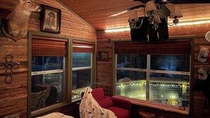 A cabin-style room with wooden walls, a ceiling fan, a mounted deer head, and a large window overlooking a lit-up bridge. - Antler Ridge Cabin One (Murfreesboro)