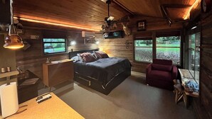 A cozy cabin-style room with a bed, a desk, and a chair. There are two windows and a mounted television. - Antler Ridge Cabin One (Murfreesboro)