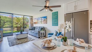 Un salon moderne comprenant un coin repas, de grandes fenêtres, un ventilateur de plafond et une cuisine équipée d’un réfrigérateur.