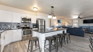 Una cocina moderna con armarios blancos, una isla central con taburetes, electrodomésticos de acero inoxidable y salpicadero de azulejos.