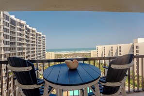 Un balcón con una mesa redonda y dos sillas, con vista a una playa y edificios altos.