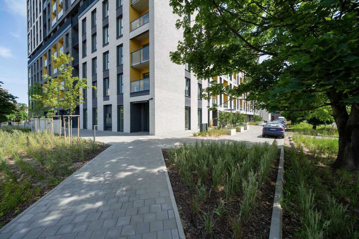 A modern apartment building with a paved walkway, a tree-lined path, and a parked car.