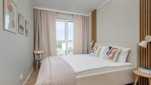 A bedroom with a bed, a window, a chair, and a bedside table with a lamp.