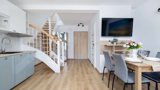 A modern kitchen with a wooden staircase, a dining area with grey chairs, and a flat-screen TV mounted on the wall.