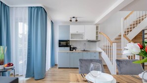 A modern kitchen with white cabinets, a microwave, and a coffee maker. There is a dining area with a table set for two, a grey sofa, and a staircase with wooden railings.