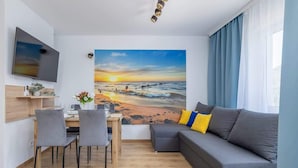 A modern living room with a grey sofa, a dining table with chairs, a flat-screen TV mounted on the wall, and a large painting of a sunset over the ocean.