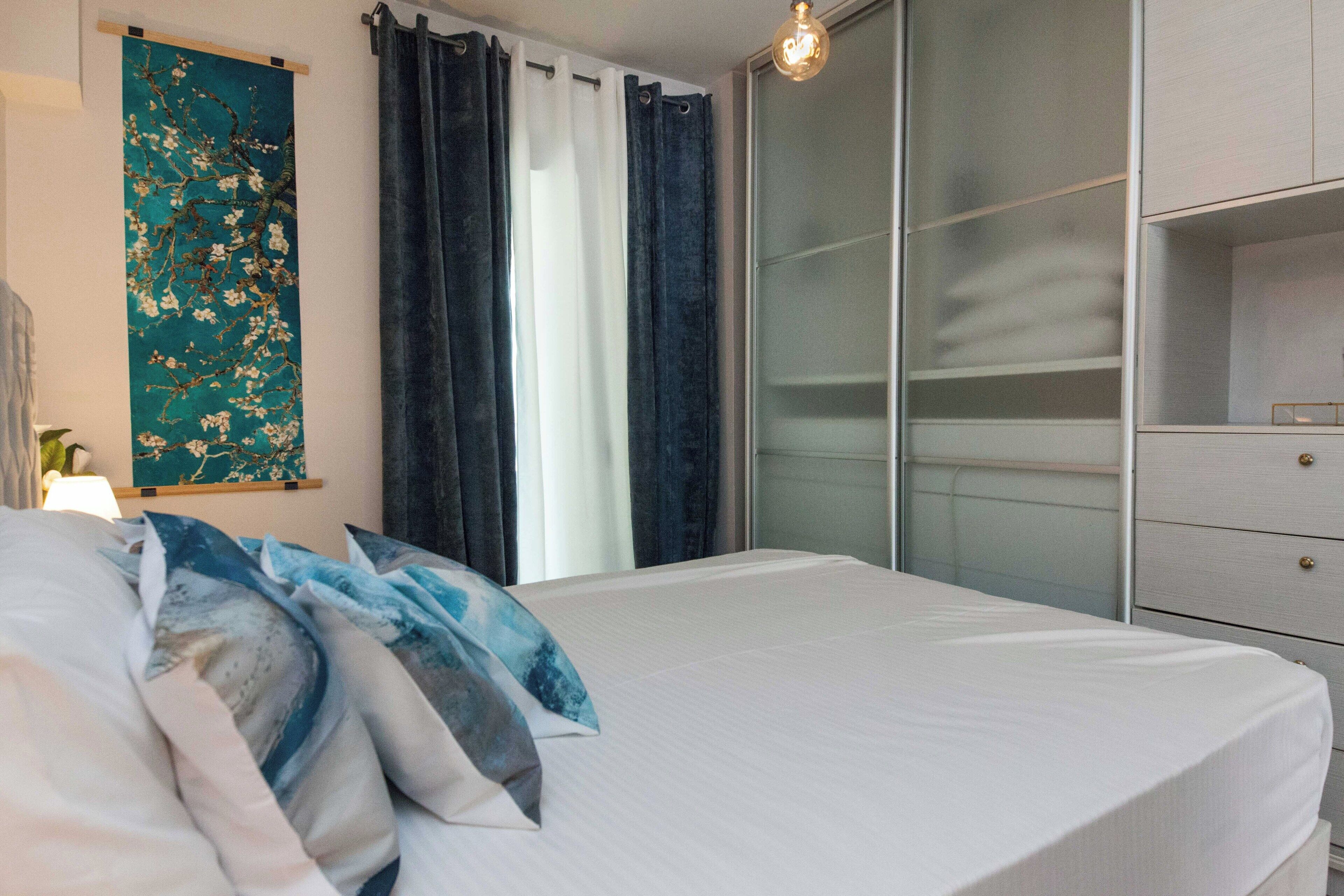 A neatly made bed with a white duvet and blue pillows, a teal floral tapestry on the wall, a dark curtain, and a white wardrobe with drawers.
