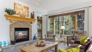 Un salon chaleureux avec une cheminée, un canapé, une table basse et une grande fenêtre donnant sur la forêt.