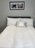 Una cama bien hecha con sábanas blancas y una única almohada de plata, frente a una pared gris con dos cuadros enmarcados encima.
