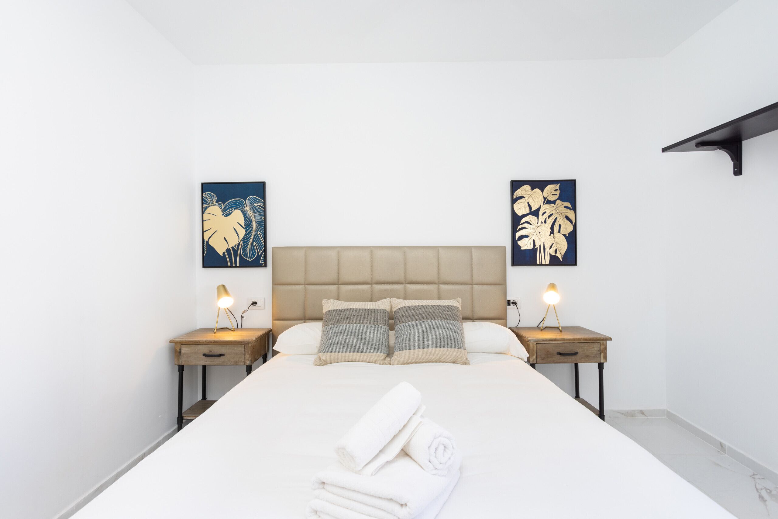 Una cama bien hecha con un cabecero color beige, dos mesitas de noche con lámparas y obras de arte en las paredes.