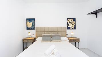 Una cama bien hecha con un cabecero color beige, dos mesitas de noche con lámparas y obras de arte en las paredes.