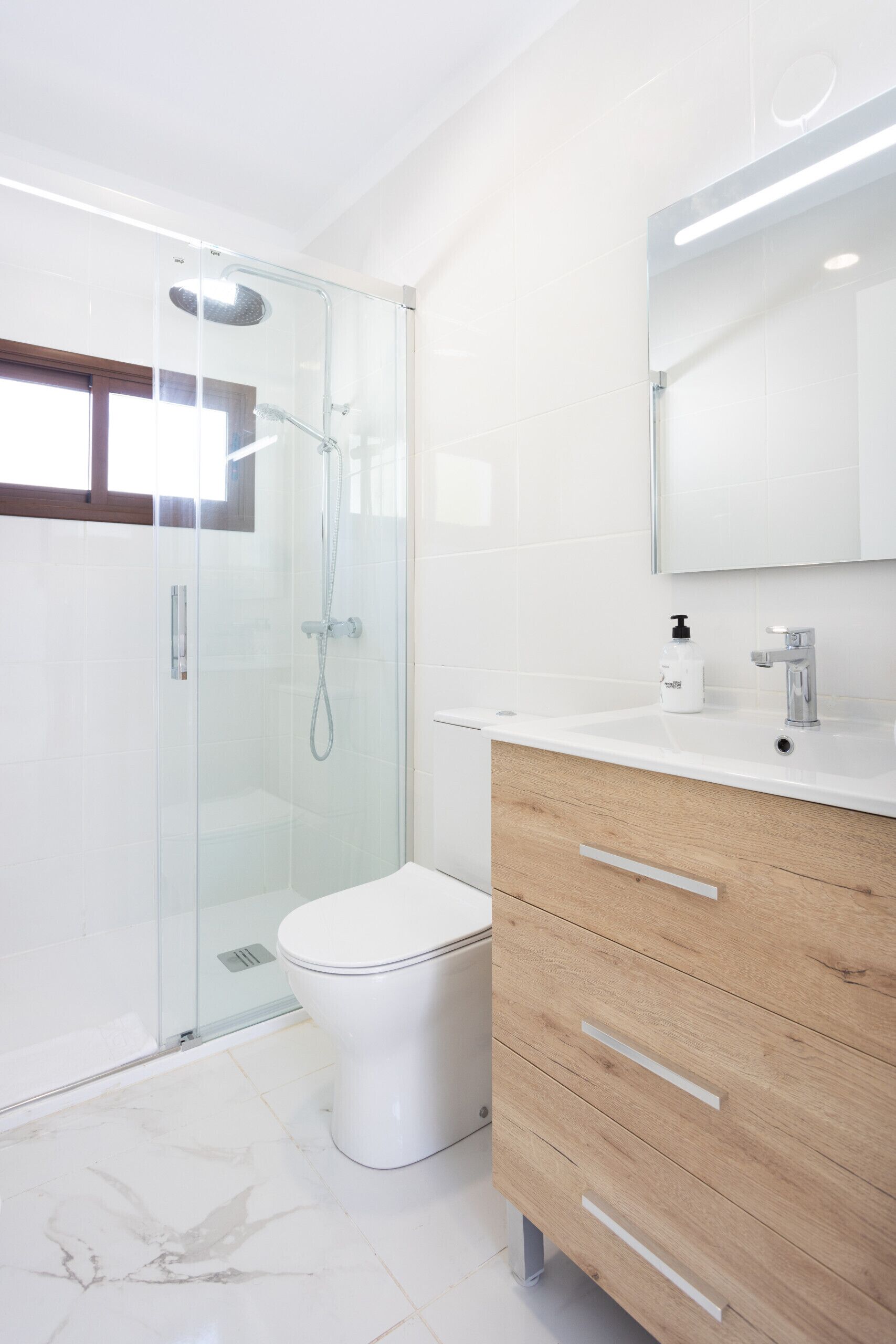 Un baño moderno con ducha envuelta en vidrio, inodoro blanco y tocador de madera con espejo.