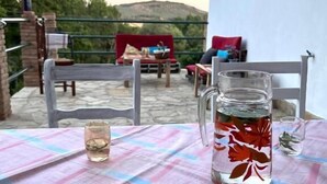 A table with a checkered tablecloth, a pitcher with leaves, and a glass of water on a balcony with chairs and a red blanket.