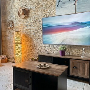 Una habitación con pared de piedra, un televisor montado, una mesa de madera, un armario y una lámpara decorativa.