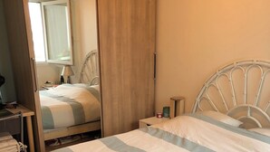 A neatly made bed with striped bedding, a wooden wardrobe, a bedside table with a lamp, and a mirror reflecting the room. - Joli Appartement T2 Vannes / 2 à 4 Personnes (Vannes)
