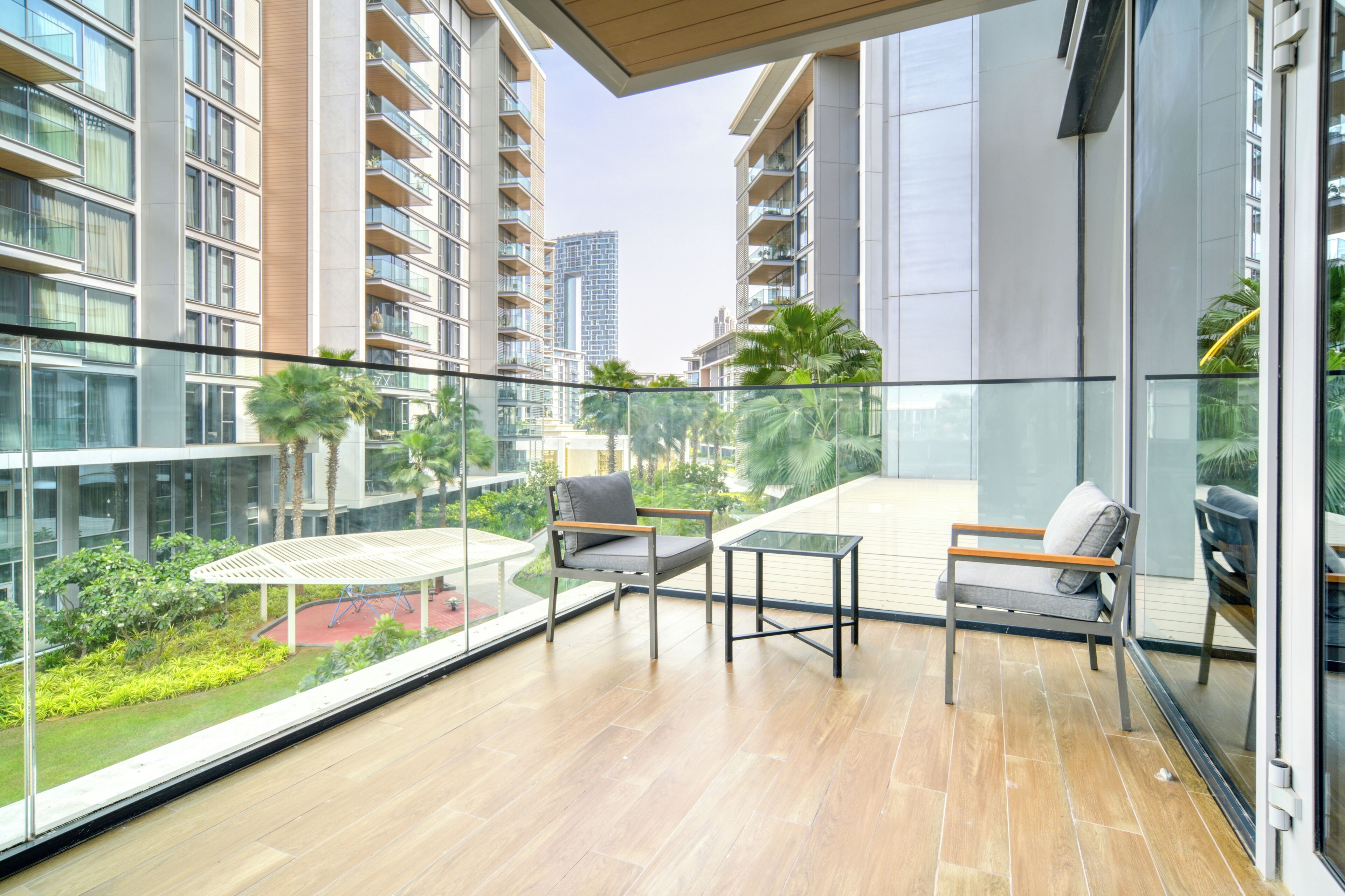 A balcony with two chairs and a small table, overlooking a lush garden and modern buildings.
