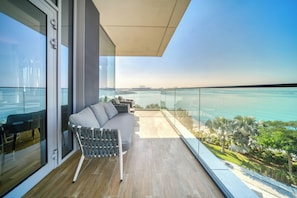 A modern balcony with a grey sofa, a glass table, and a view of the sea and city skyline.