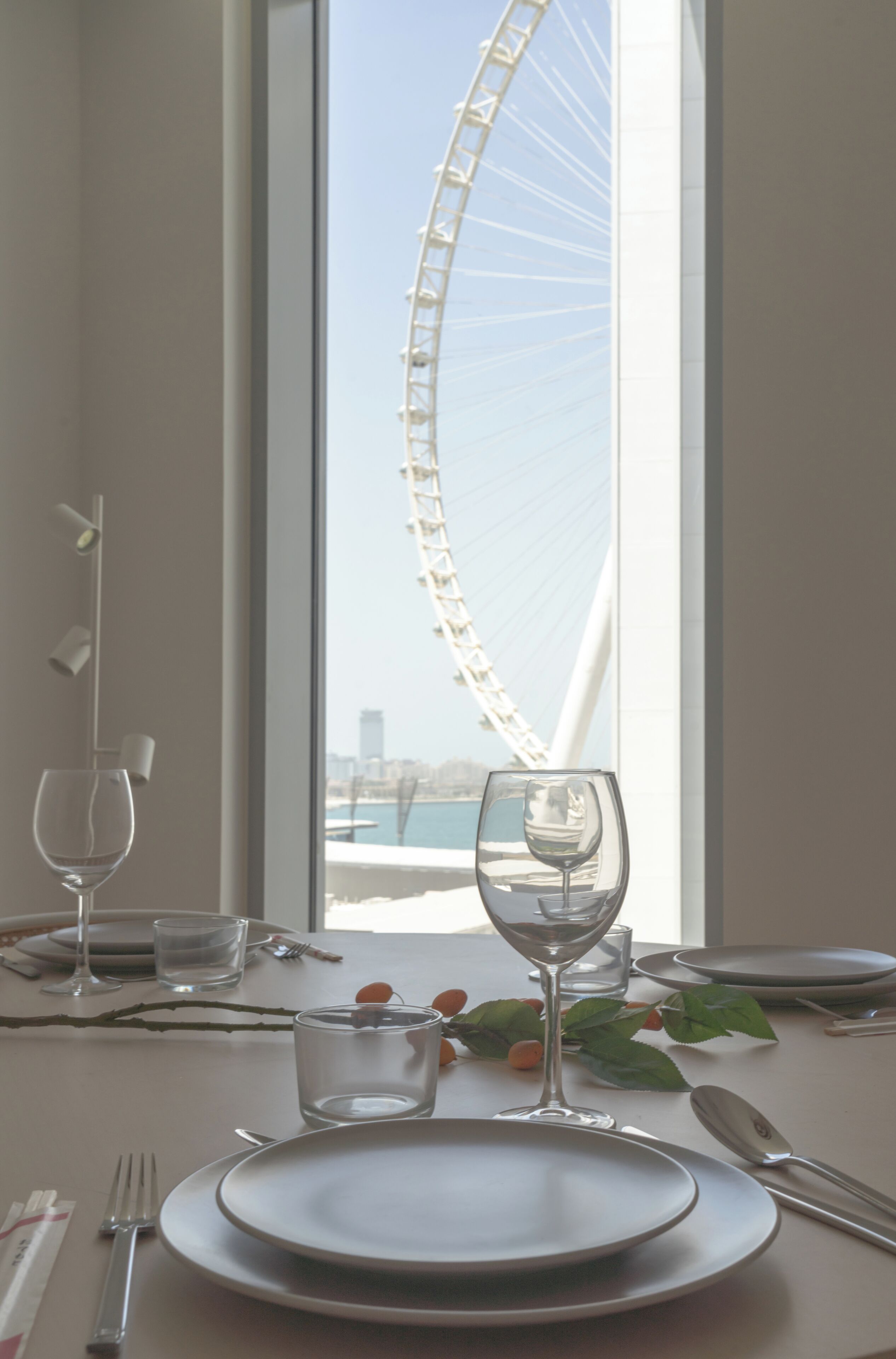 A dining table set with plates, cutlery, and wine glasses, with a view of a large Ferris wheel through a window.