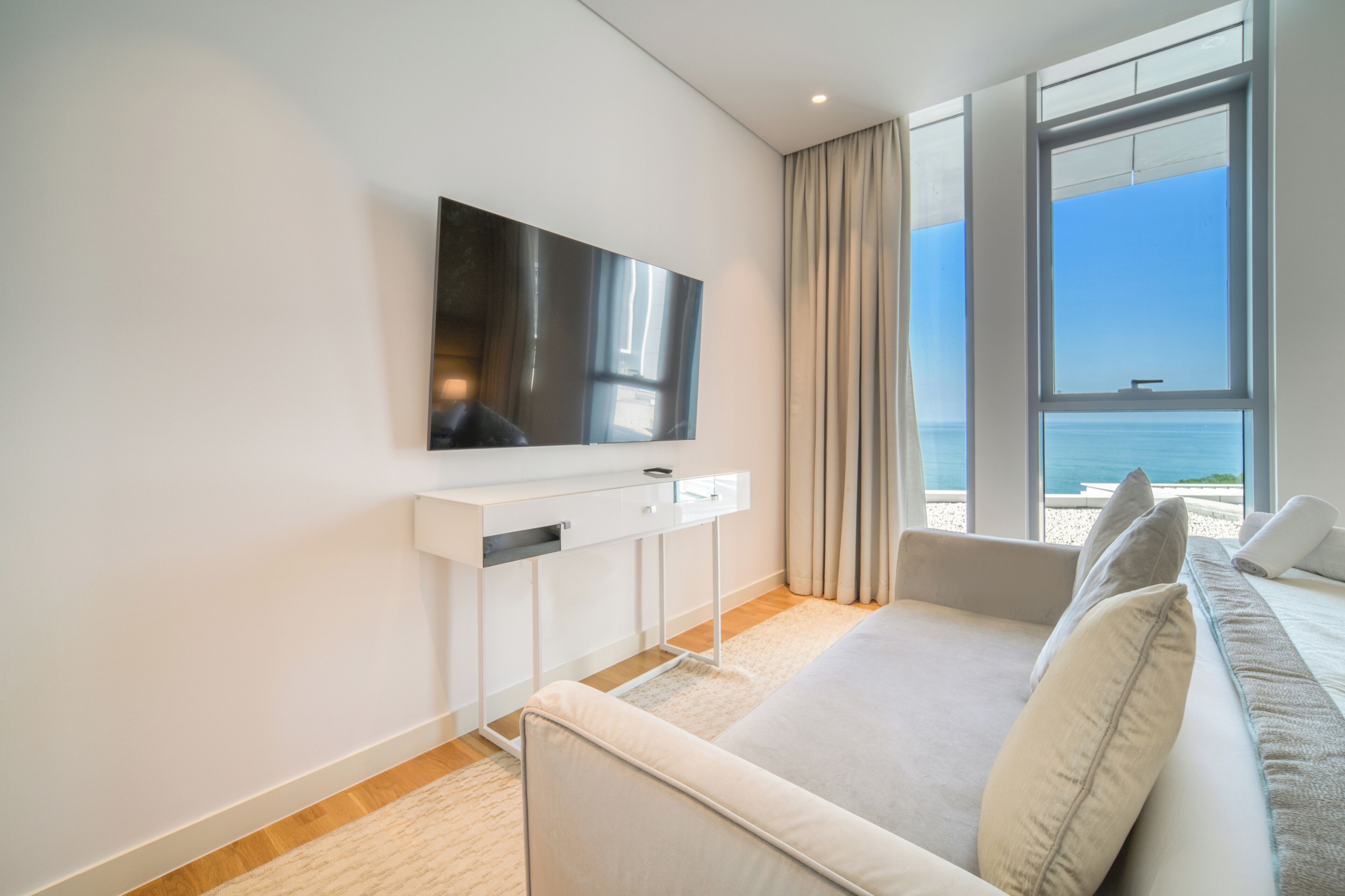 A modern hotel room with a large flat-screen TV mounted on the wall, a white console table, a beige sofa, and a large window offering a view of the sea.
