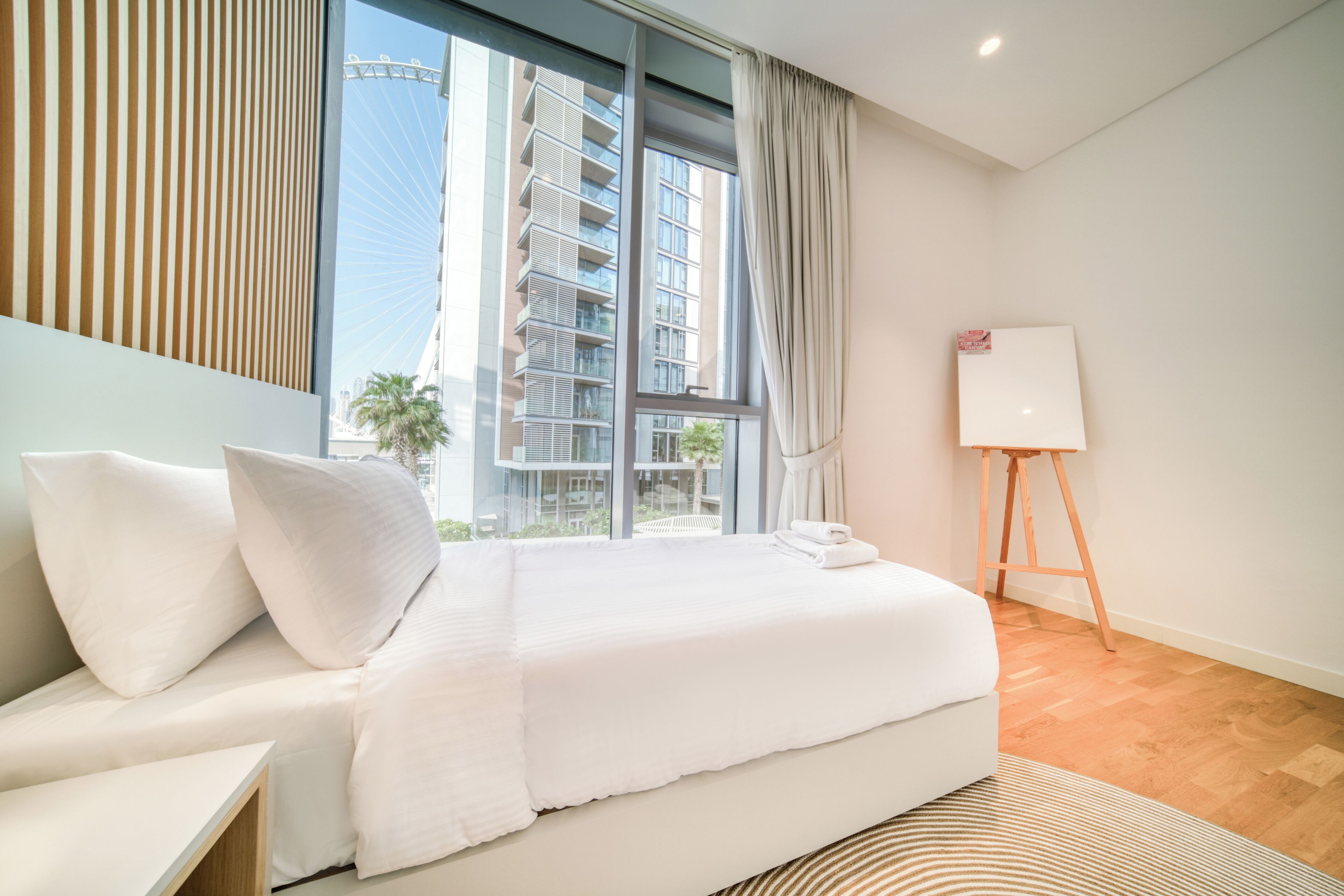 A modern hotel room with a large bed, a bedside table, a whiteboard, and a view of a tall building.