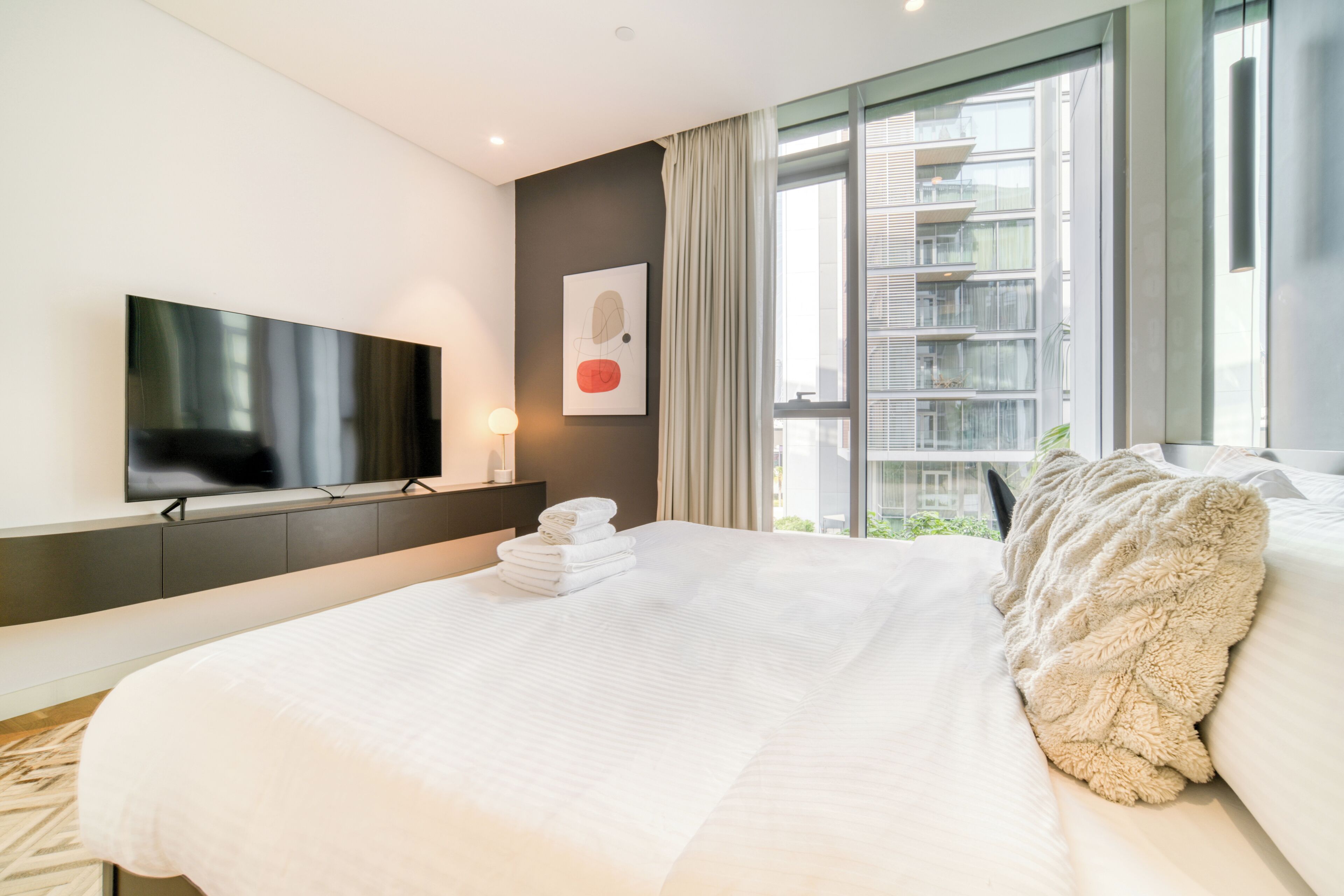 A modern hotel room with a large bed, a flat-screen TV mounted on the wall, a window with a view of a building, and a neatly made bed with a fluffy pillow.