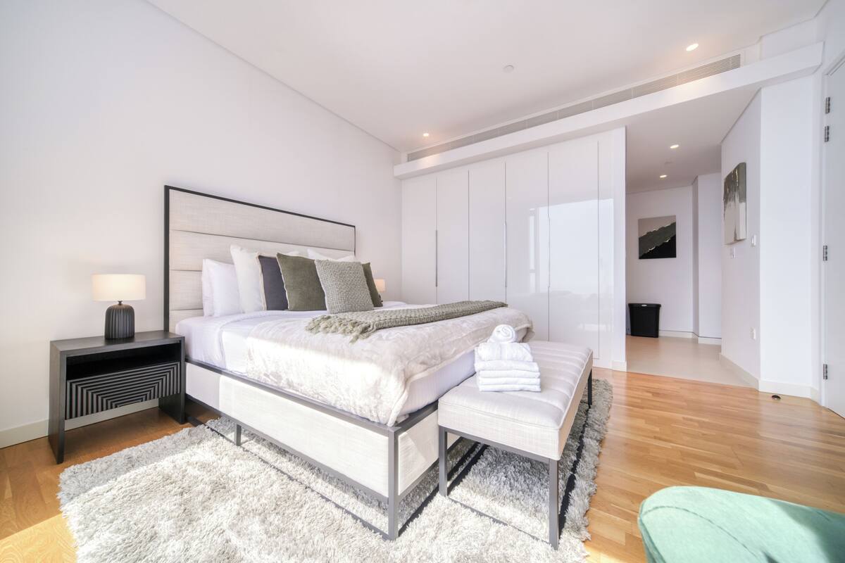 A modern bedroom with a large bed, bedside tables, a bench, and a walk-in closet.