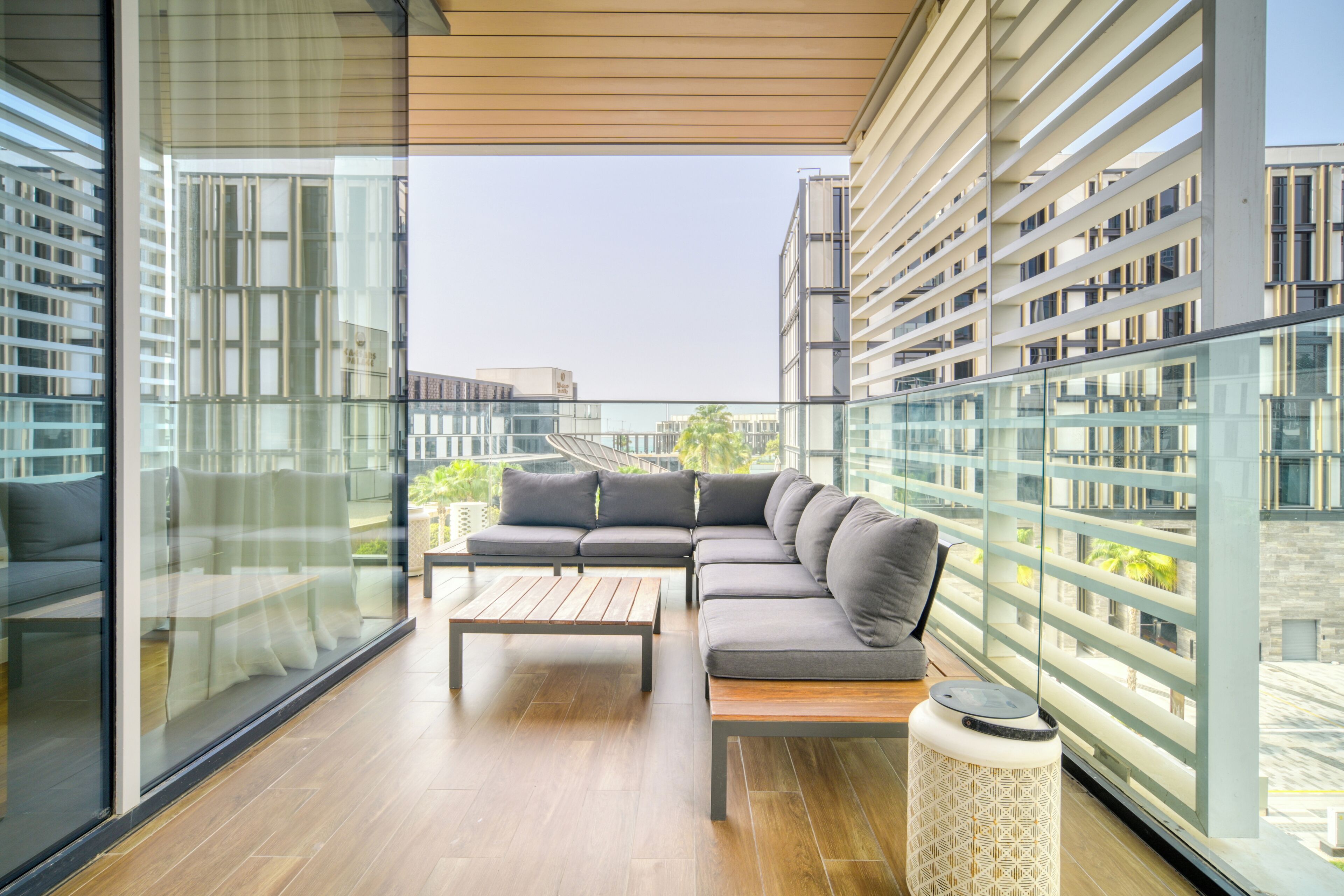 A modern balcony with a sectional sofa, a wooden coffee table, and a patterned ottoman.