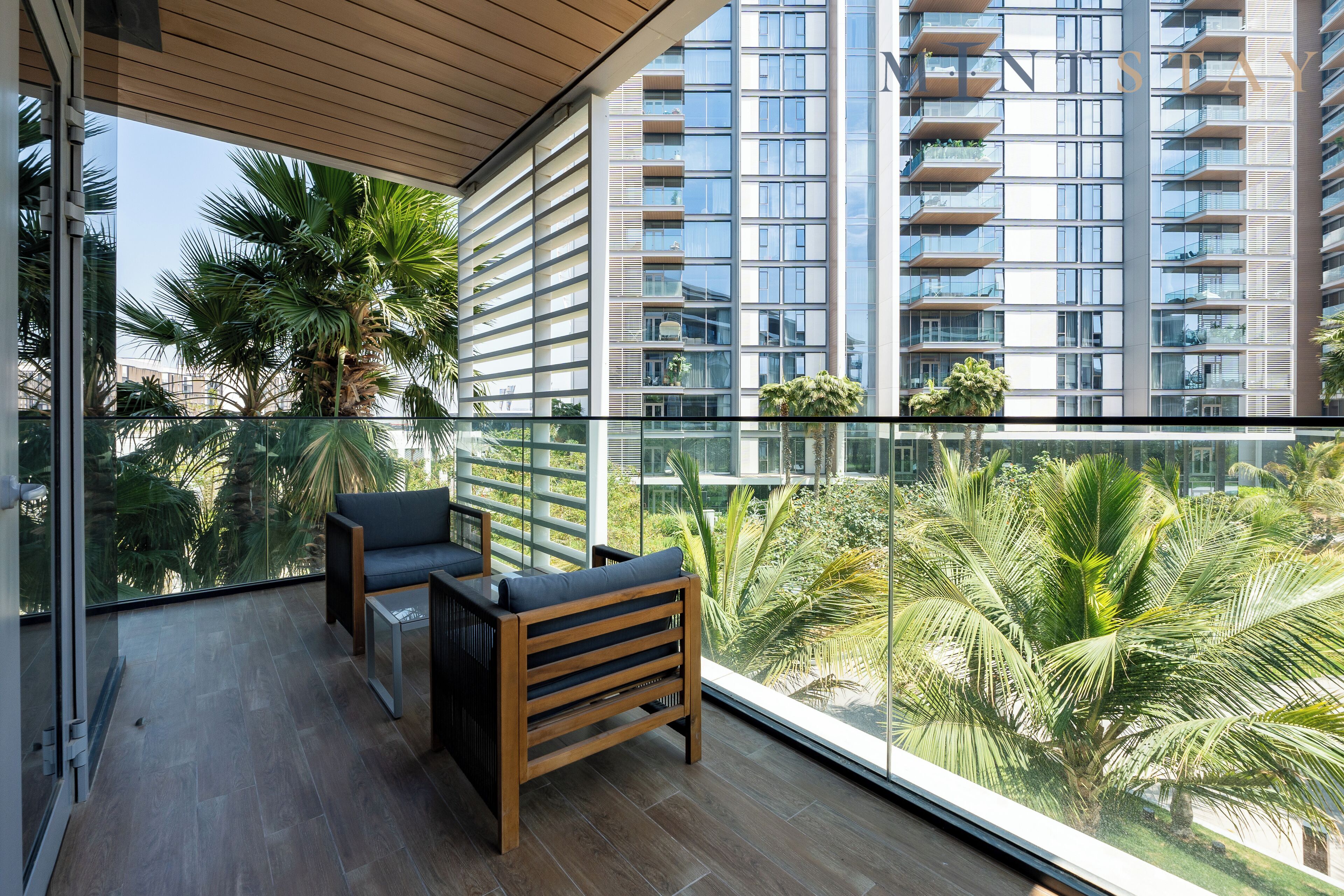 A balcony with a wooden bench and a glass table, overlooking a cityscape with tall buildings and palm trees.