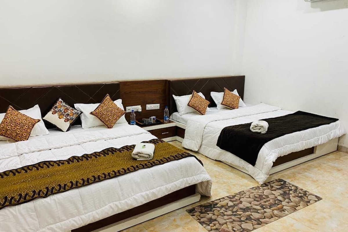 A hotel room with two beds, each with white bedding and decorative pillows. There is a small table with toiletries and a water bottle. The room has a tiled floor and a decorative rug.