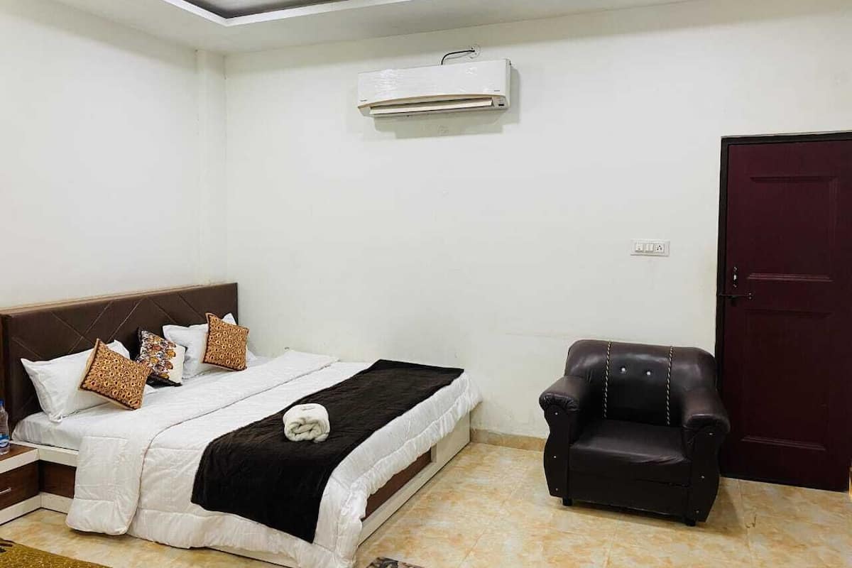 A bedroom with a bed, a black leather armchair, a dark wooden door, and an air conditioning unit on the wall.