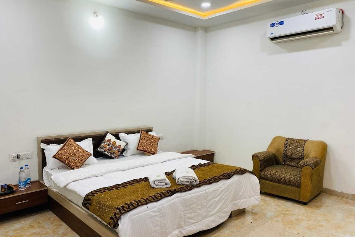 A hotel room with a bed, a chair, an air conditioning unit, and a nightstand with water bottles.
