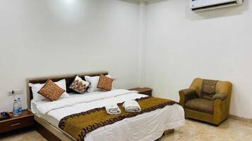 A hotel room with a bed, a chair, an air conditioning unit, and a nightstand with water bottles.
