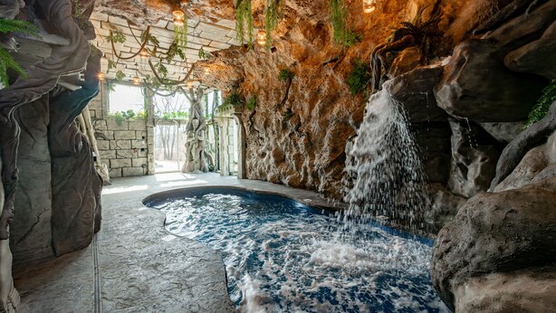 A spa area with a hot tub, a waterfall feature, and a stone wall with greenery. - New! Epic lakefront home with indoor pool! (McHenry)
