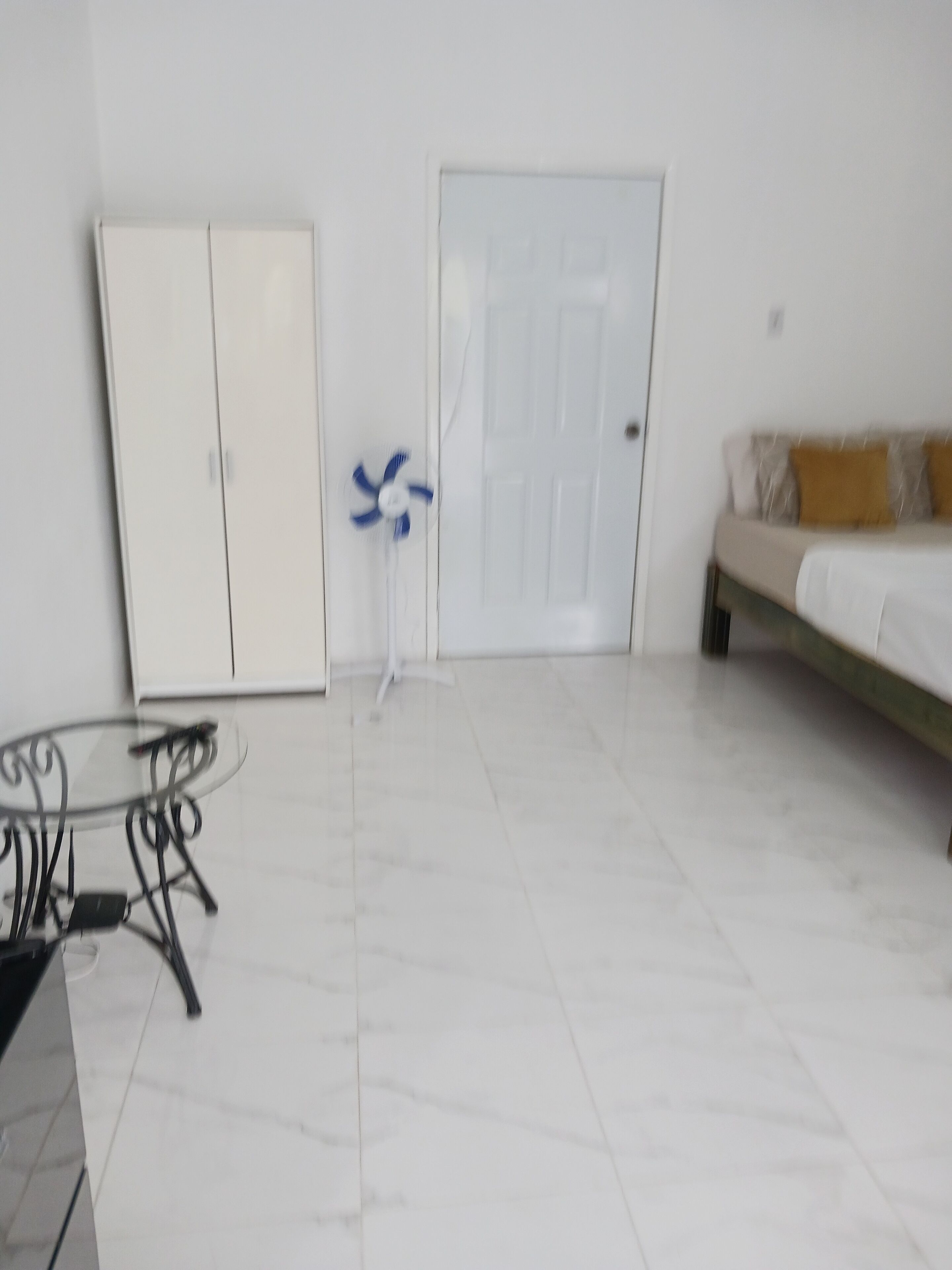 A room with a white wardrobe, a closed door, a small table with a fan, and a bed with a beige and brown pillow.