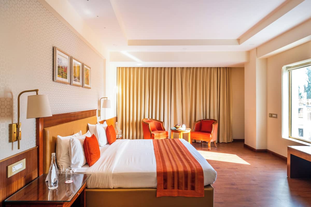 Quarto de hotel com cama grande, duas cadeiras, televisão e janela com cortinas.