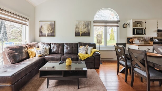 A modern living room with a leather sofa, a glass coffee table, and a dining area with wooden chairs. There are two large windows providing natural light and a view of the outdoors. - Cozy and close to beach ,3 bedroom condo in Magog (Magog)