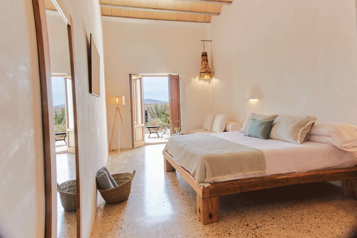 Quarto com cama de madeira, roupa de cama branca, cesto de vime e vista para o exterior.