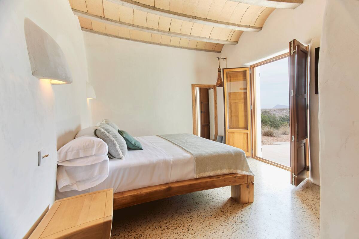 Quarto com cama de madeira, mesa de cabeceira, luminária na parede e uma janela grande com vista para o exterior.