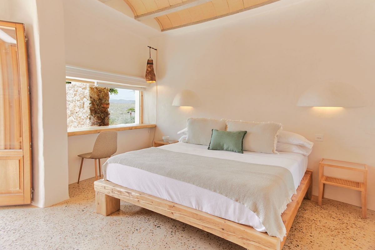 Quarto com cama de madeira, janela com vista, uma cadeira e mesa de cabeceira.
