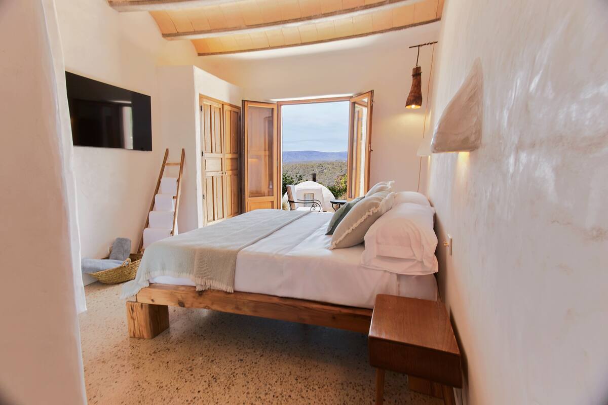 Quarto com cama de madeira, vista do deserto por uma porta aberta e uma mesa pequena com luminária.
