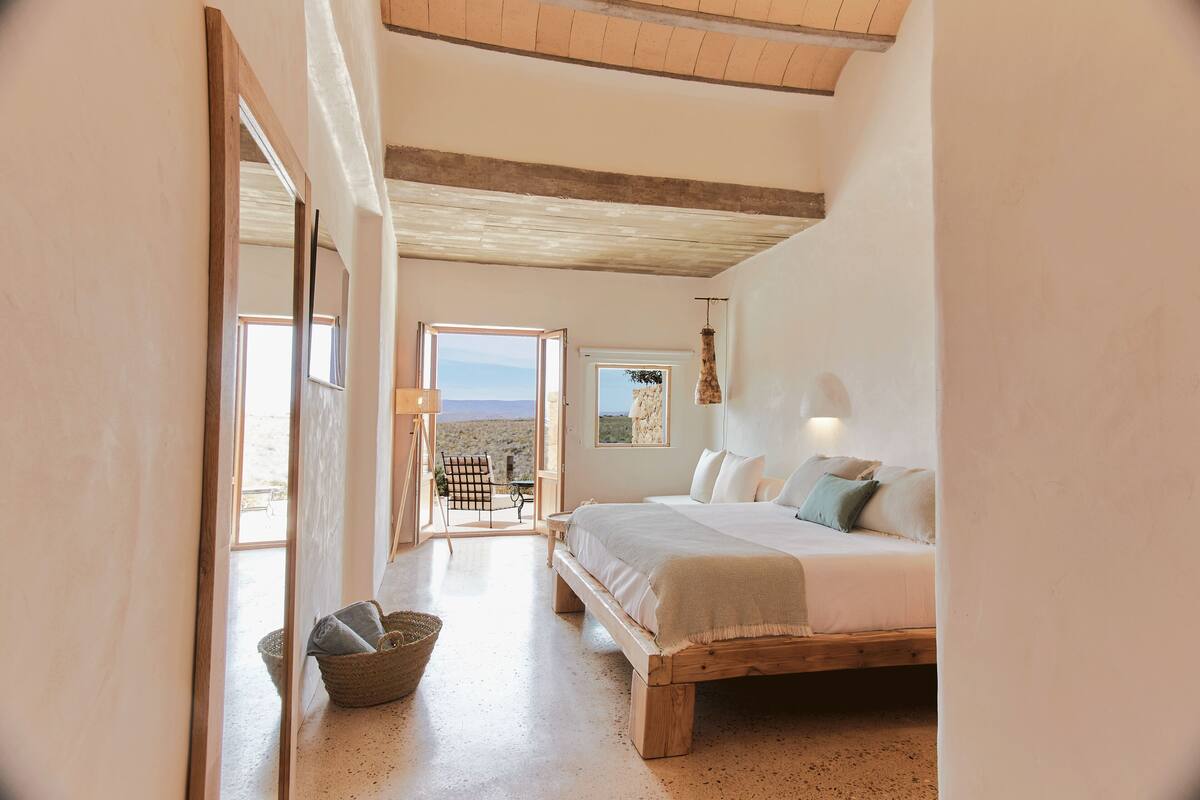 Quarto com uma cama grande, um banco de madeira, um cesto de vime e vista para o exterior.