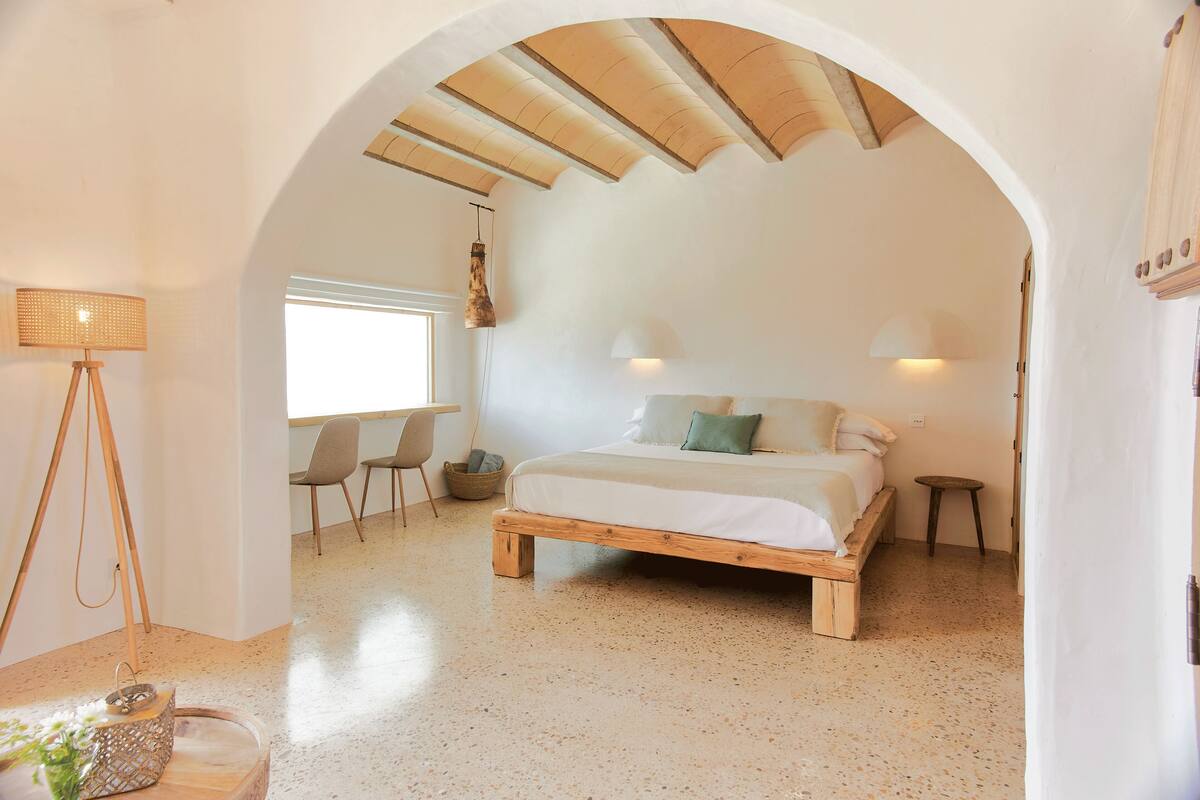 Quarto com cama de madeira, escrivaninha e cadeira, luminária pendente e janela.