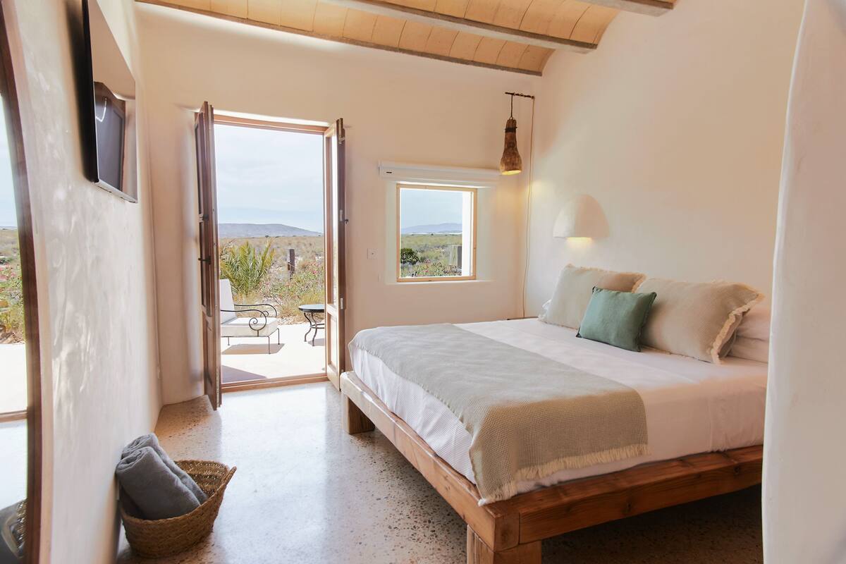 Quarto com cama de madeira, pequena janela, luminária na parede e vista para o exterior.