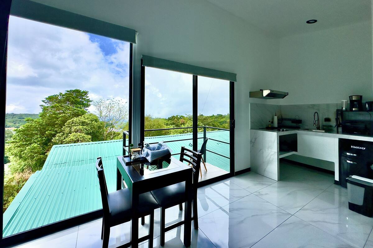 Une cuisine moderne dotée d’une grande fenêtre, un coin repas avec une table et des chaises, et une vue sur la verdure à l’extérieur.