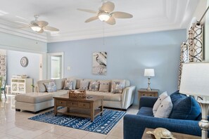 A spacious living room with a beige sectional sofa, a blue armchair, a wooden coffee table, and a blue patterned rug. There are two ceiling fans, a wall-mounted lamp, and a framed artwork above the sofa.