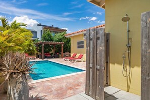 A swimming pool with a wooden pergola, red lounge chairs, and a wooden door leading to a shower area.